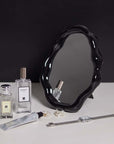 Nordic cloud-shaped vanity mirror with a black wavy frame on a table alongside perfumes, jewelry, and a watch.