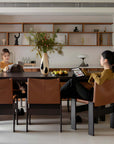 Elegant dining room with Gora dining table, cherry wood, Monk chairs with leather seats, women enjoying cozy ambiance, modern decor.