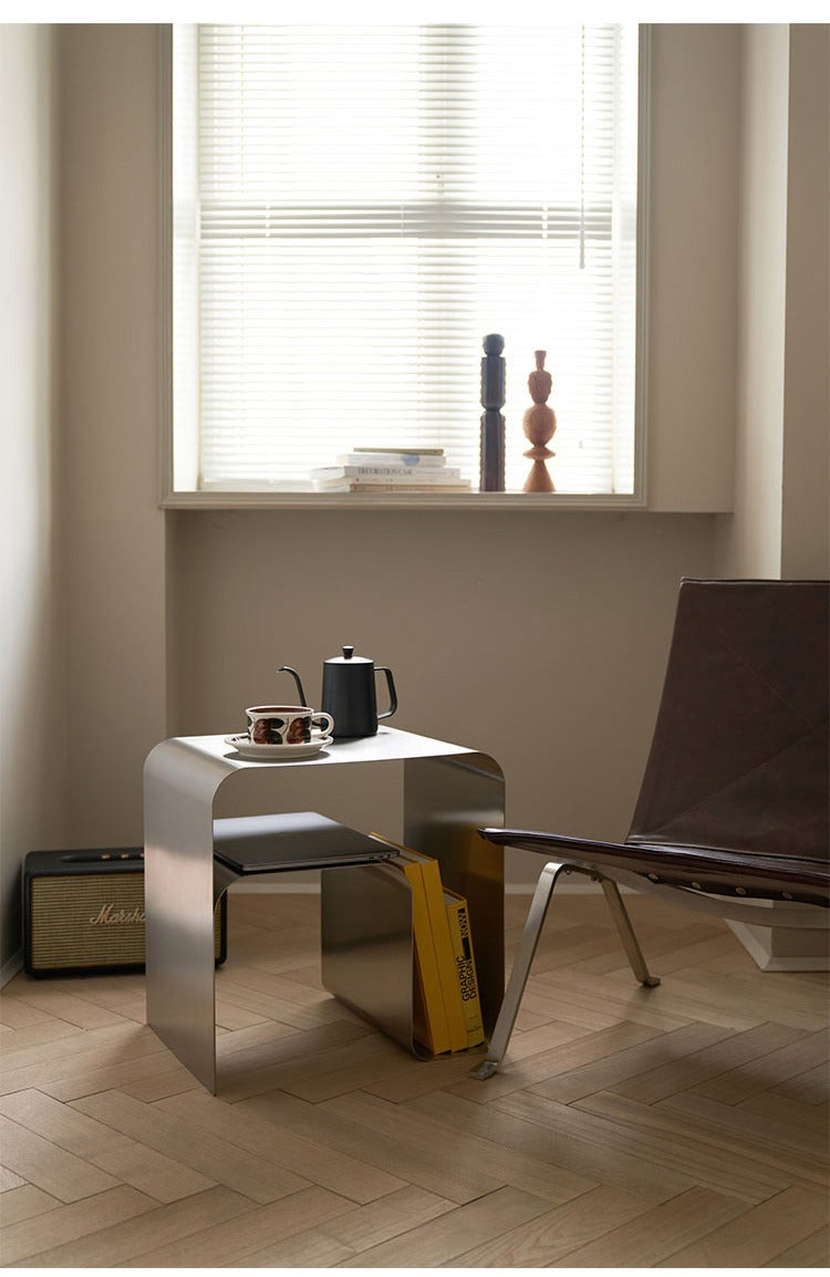 Minimalist stainless steel bedside table with books and a coffee set in a modern room setting.