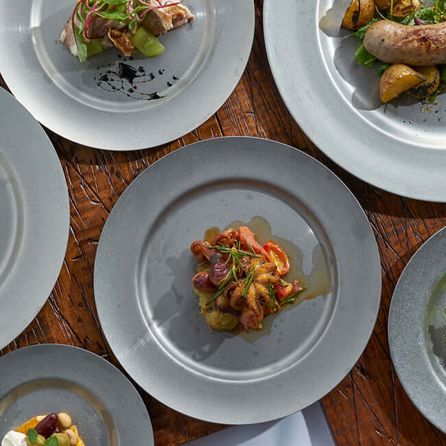 Stainless steel round plates with gourmet dishes on a wooden table, showcasing VINTAGE INOX’s chic design and durability.