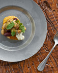 Vintage INOX round stainless steel plate with gourmet appetizer on rustic wooden table with spoon.
