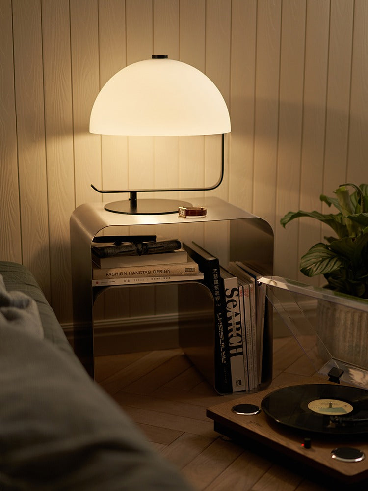 Minimalist stainless steel bedside table with a modern lamp, books, and a cozy living room setting.