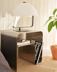 Minimalist stainless steel bedside table with lamp, books, and magazine in a cozy room setting with plant decor.