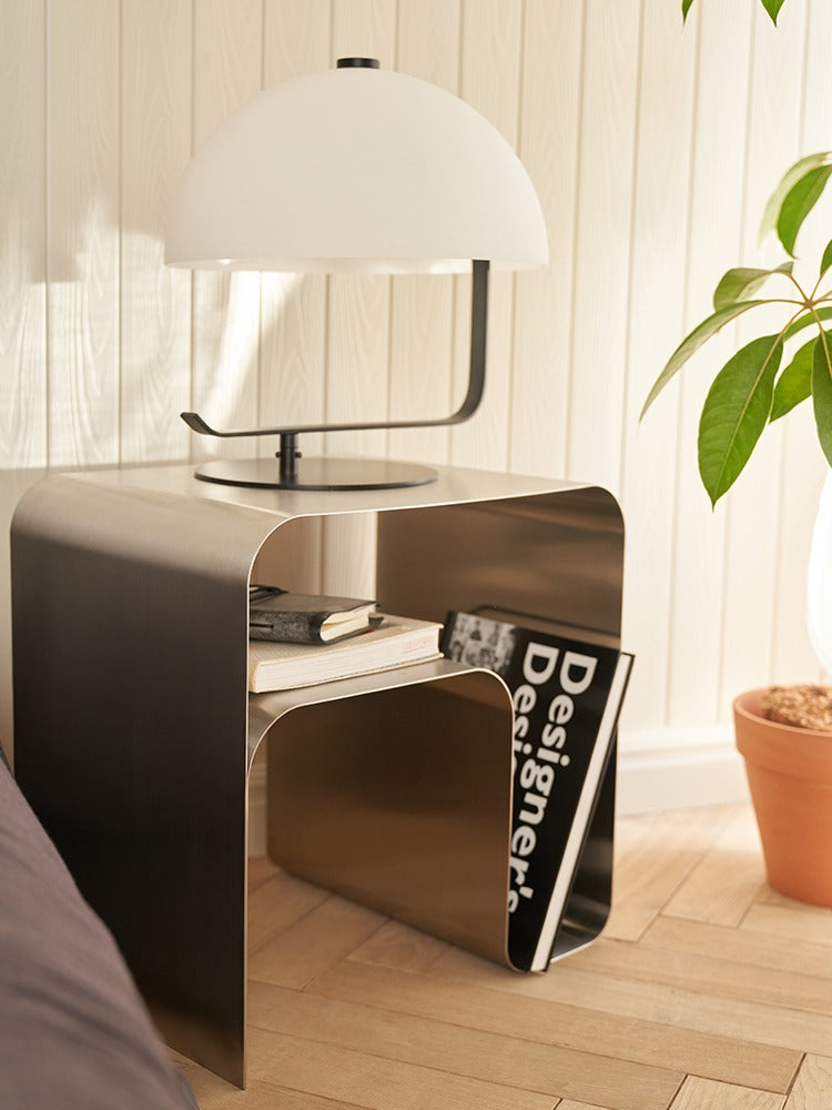 Minimalist stainless steel bedside table with lamp, books, and magazine in a cozy room setting with plant decor.