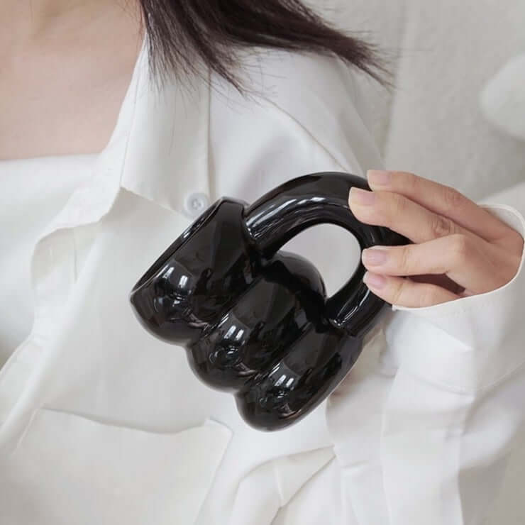 Person holding a black ceramic mug shaped like brass knuckles, wearing a white shirt.