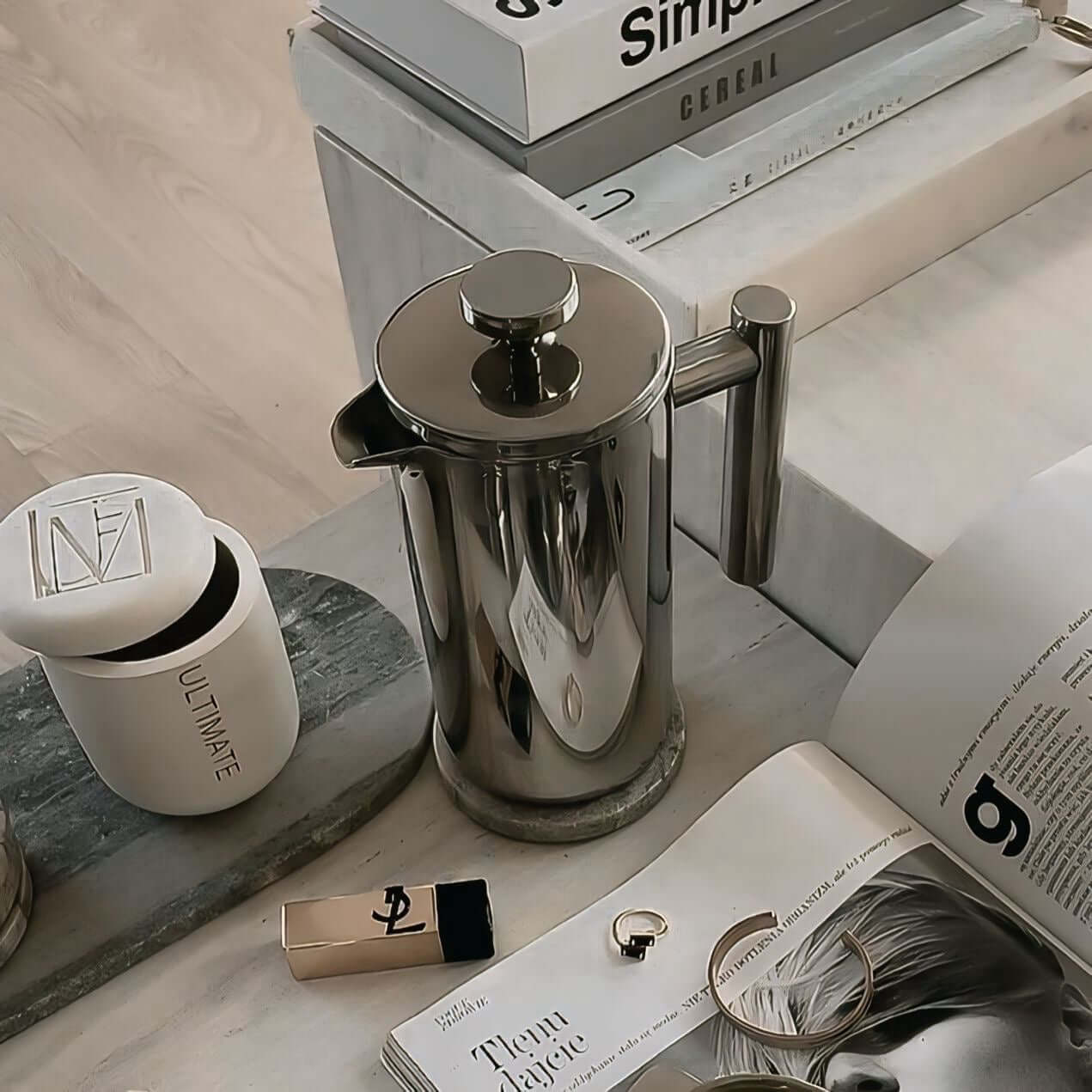 Stainless steel French press coffee pot with mirror finish on a table surrounded by elegant accessories and a magazine.