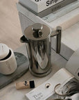 Stainless steel French press coffee pot with mirror finish on a table surrounded by elegant accessories and a magazine.