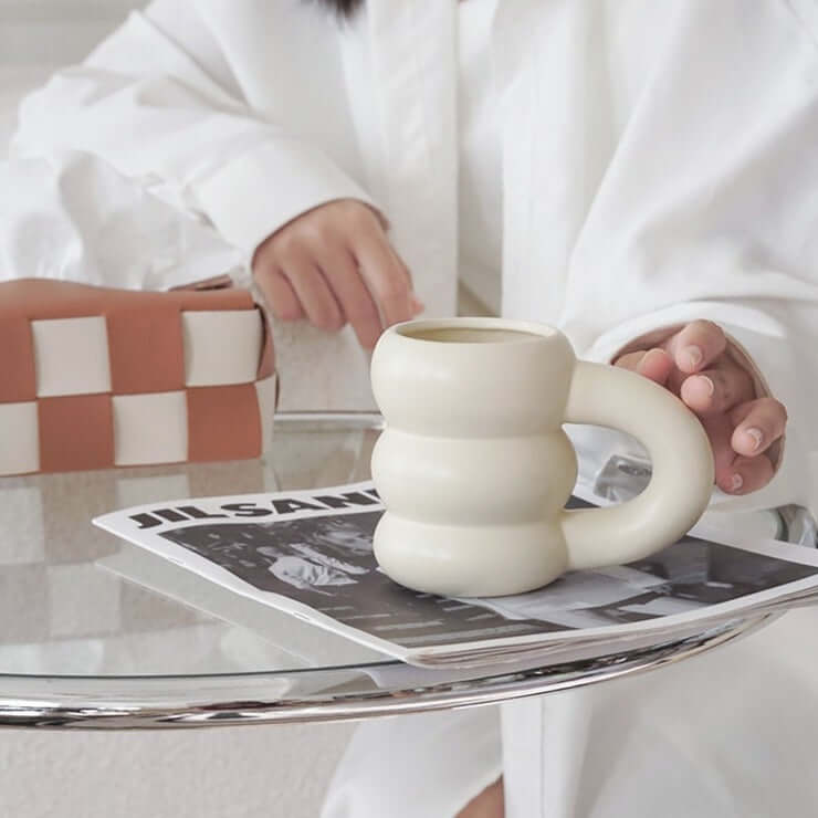 Unique cream-colored ceramic mug with a donut shape, placed on a glass table next to a magazine in a stylish setting.