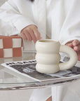 Unique cream-colored ceramic mug with a donut shape, placed on a glass table next to a magazine in a stylish setting.