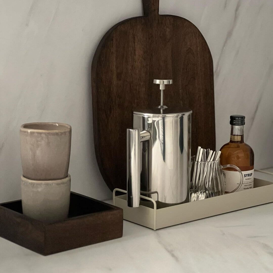 Stainless steel French press coffee pot on a tray with cups and syrup on a kitchen counter setup.