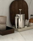 Stainless steel French press coffee pot with mirror finish on kitchen counter with cups and bottle.