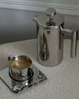 Stainless steel French press coffee pot with double-layer insulation next to a cup of coffee on a stylish reflective tray.