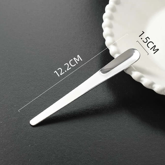 Minimalist dessert spoon with measurements of 12.2cm length and 1.5cm width displayed beside a scalloped plate.