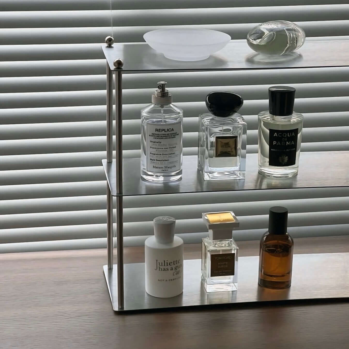 Modern Aira stainless shelf displaying perfume bottles near window blinds.