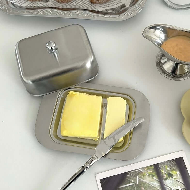 Stainless steel butter dish with lid, featuring two butter sticks on a breakfast table setting.