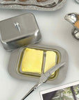 Stainless steel butter dish with lid, featuring two butter sticks on a breakfast table setting.