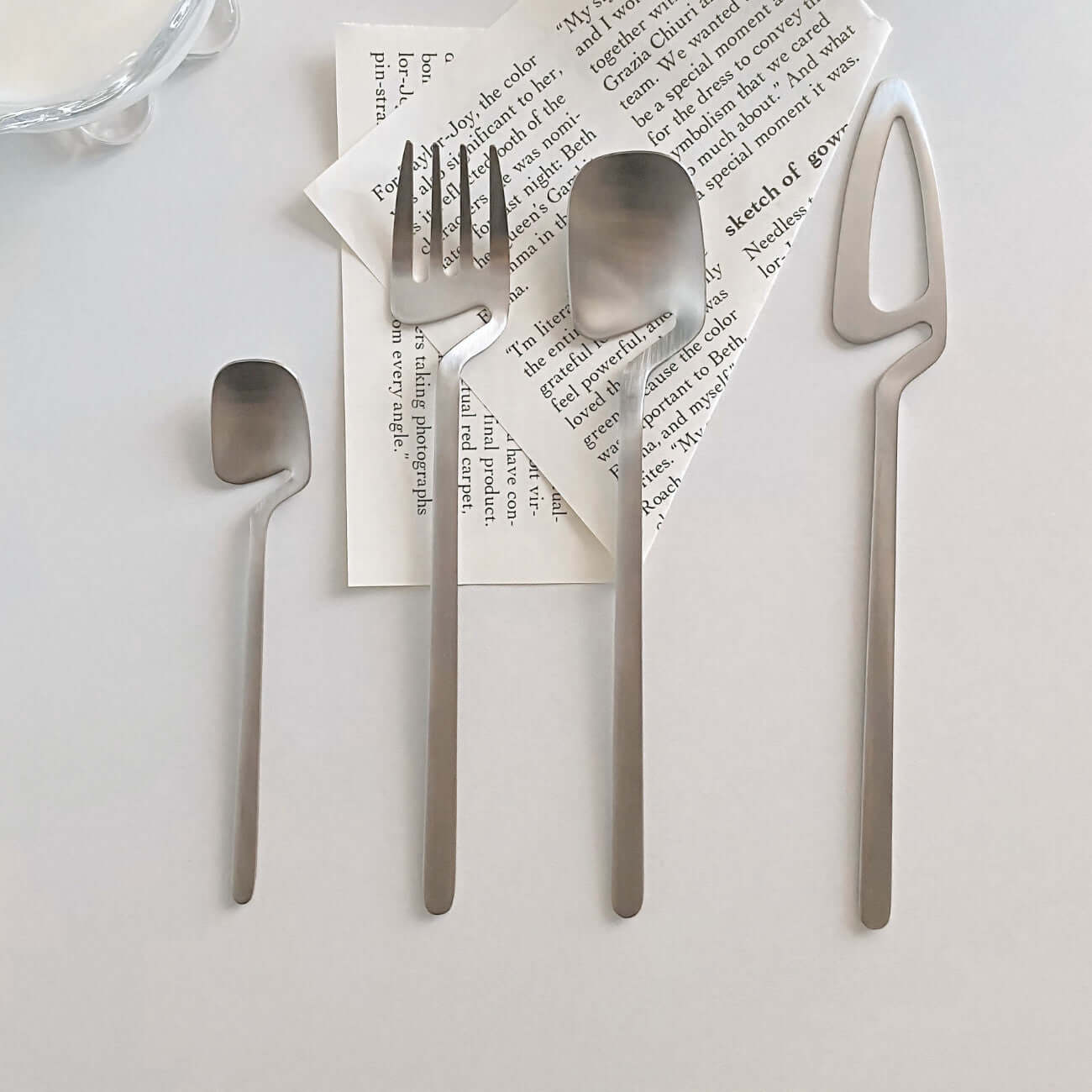 Minimalist stainless steel flatware set with small spoon, fork, spoon, and cheese slicer on a modern table setting.