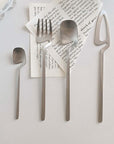 Minimalist stainless steel flatware set with small spoon, fork, spoon, and cheese slicer on a modern table setting.