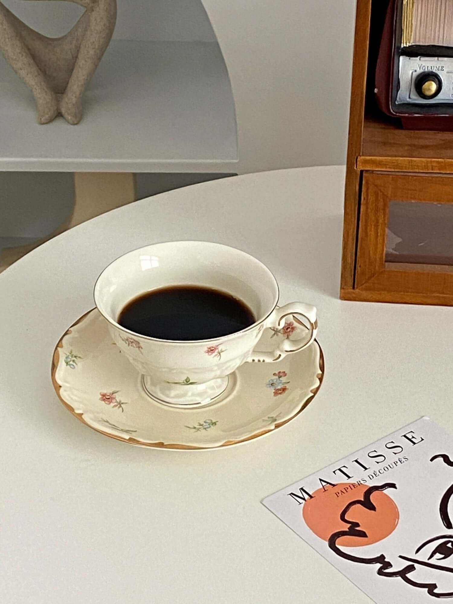 Retro French blossom cup and saucer set with coffee, placed on a white table near a book and wooden shelf.