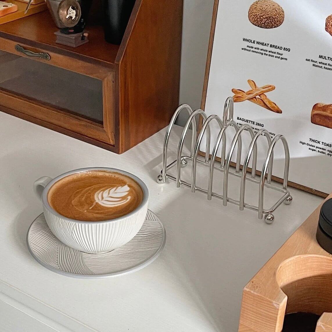 Stainless steel toast holder next to a cappuccino on a table, perfect for keeping toast warm and crisp at breakfast.