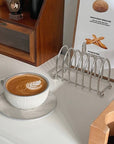 Stainless steel toast holder next to a cappuccino on a table, perfect for keeping toast warm and crisp at breakfast.