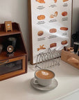 Stainless steel toast holder on a kitchen counter with coffee and bread chart in background.