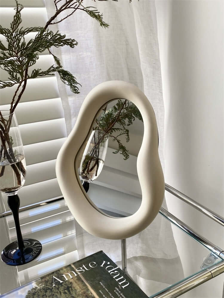 Cream Nordic asymmetric mirror on a glass table, 29x22 inches, reflecting a plant. Free shipping available. Modern home decor accessory.