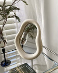 Cream Nordic asymmetric mirror on a glass table, 29x22 inches, reflecting a plant. Free shipping available. Modern home decor accessory.