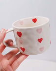 Hand-painted beige coffee mug with red and pink hearts design, 300ml capacity, shown in hand against a light background.