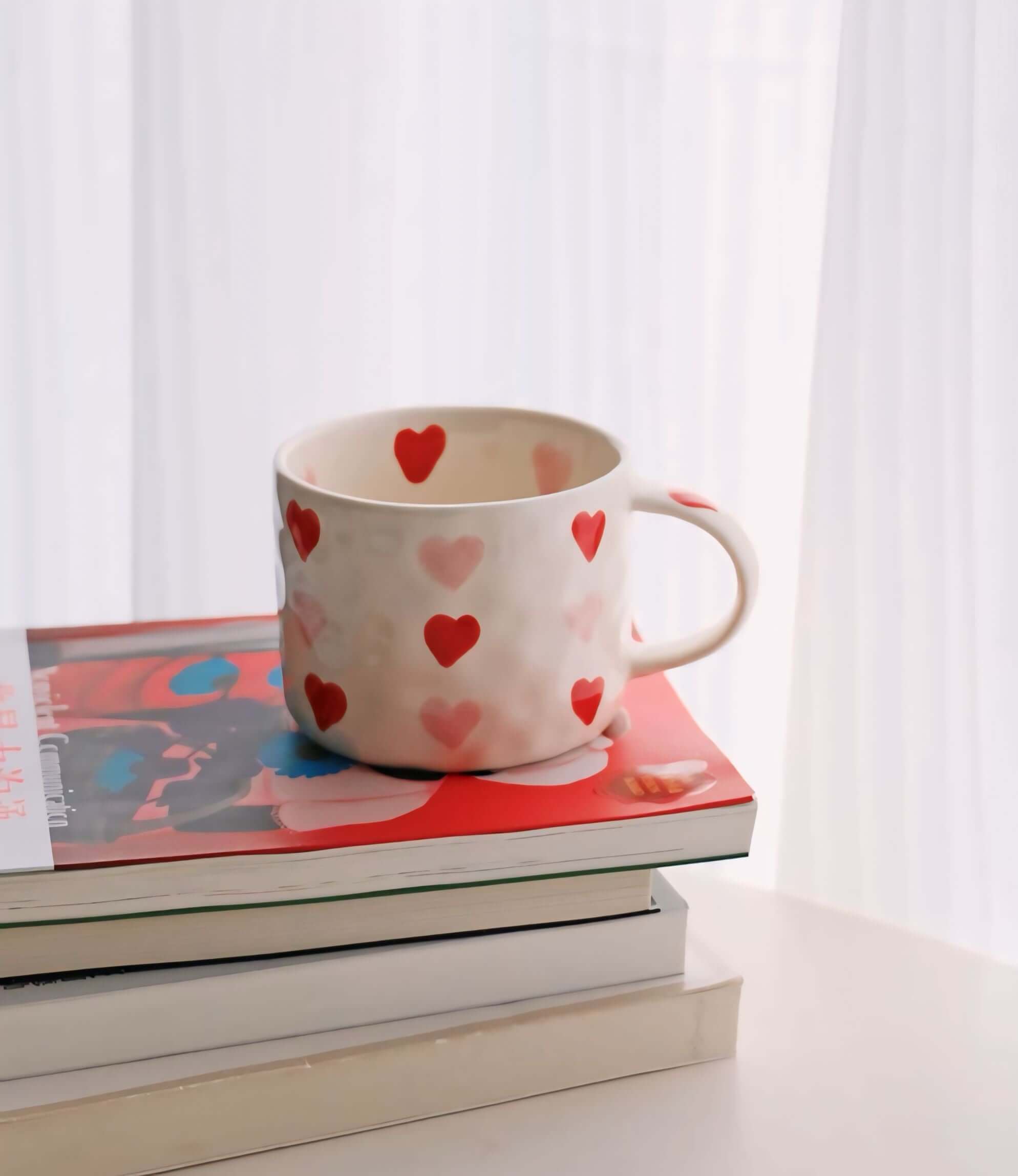 Hand-painted coffee mug with red and pink hearts, 300ml capacity, beige ceramic, placed on stacked books near a window.