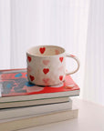 Hand-painted coffee mug with red and pink hearts, 300ml capacity, beige ceramic, placed on stacked books near a window.