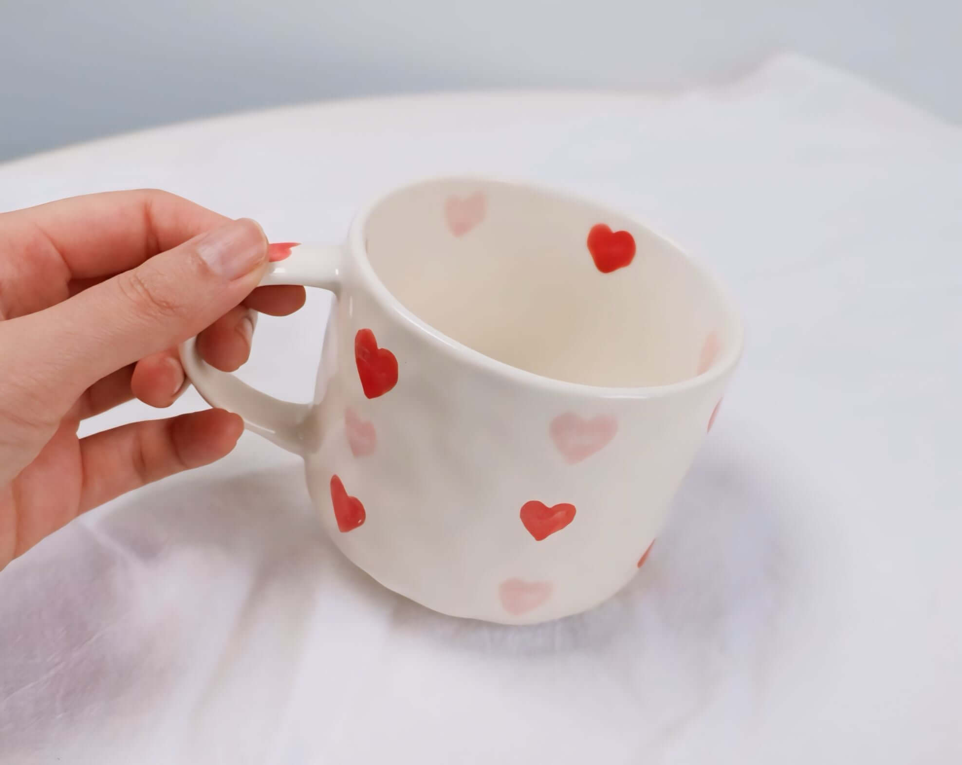 Hand holding a beige hand-painted coffee mug with red and pink heart designs, showcasing its handcrafted charm.