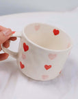 Hand holding a beige hand-painted coffee mug with red and pink heart designs, showcasing its handcrafted charm.