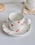 White ceramic mug and saucer set with red heart designs on a table.