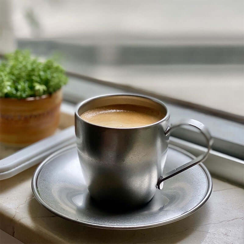 Stainless steel vintage inox cup and saucer with coffee on a windowsill, greenery in background.