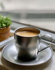 Stainless steel vintage inox cup and saucer with coffee on a windowsill, greenery in background.
