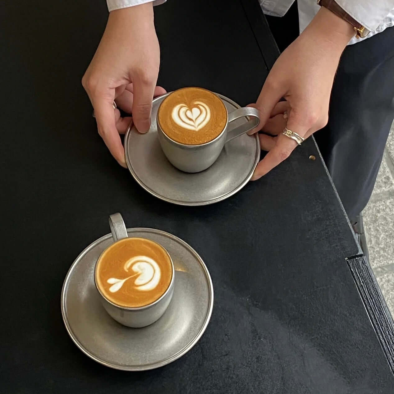 Hands placing stainless steel cups with latte art on saucers on a dark table.