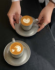 Hands placing stainless steel cups with latte art on saucers on a dark table.
