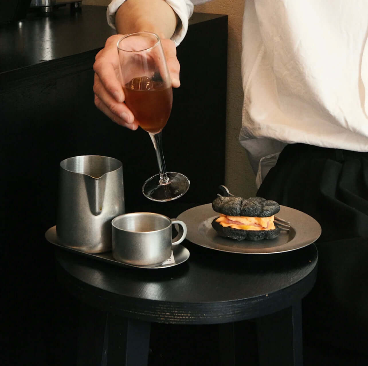 Person enjoying a drink with a stainless steel tea set and dessert on a modern black table.