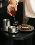 Person enjoying a drink with a stainless steel tea set and dessert on a modern black table.