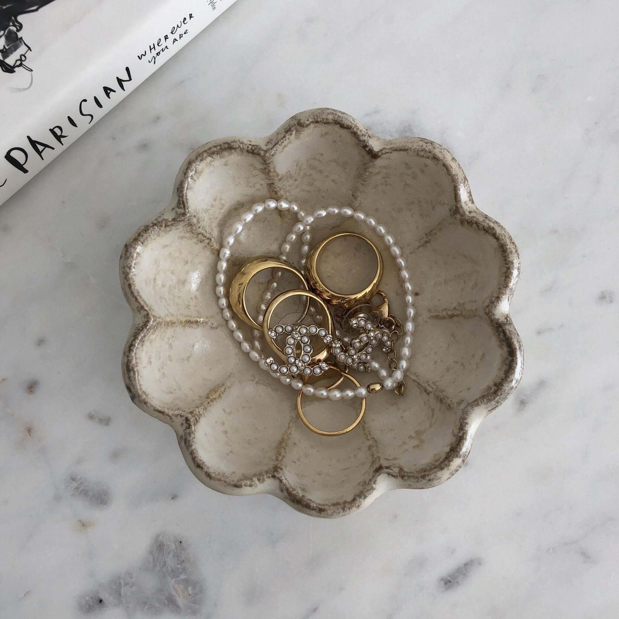Ceramic scalloped dish with gold rings and a pearl necklace on a marble surface.
