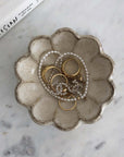 Ceramic scalloped dish with gold rings and a pearl necklace on a marble surface.