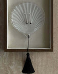 Lumière Shell Glass Dish with a black tassel on a travertine surface, framed