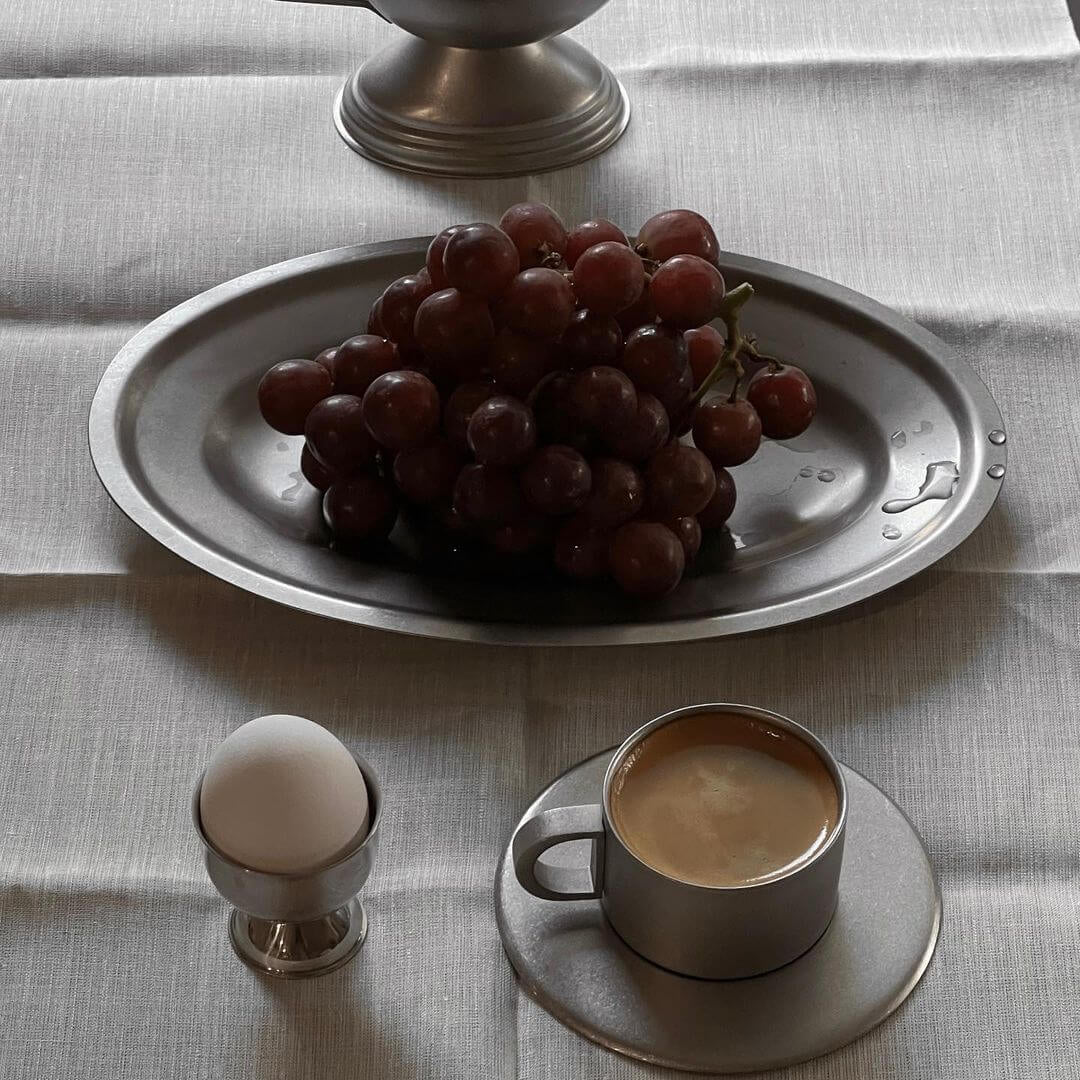 Stainless steel oval plate with grapes, egg holder, and coffee cup on a linen tablecloth.