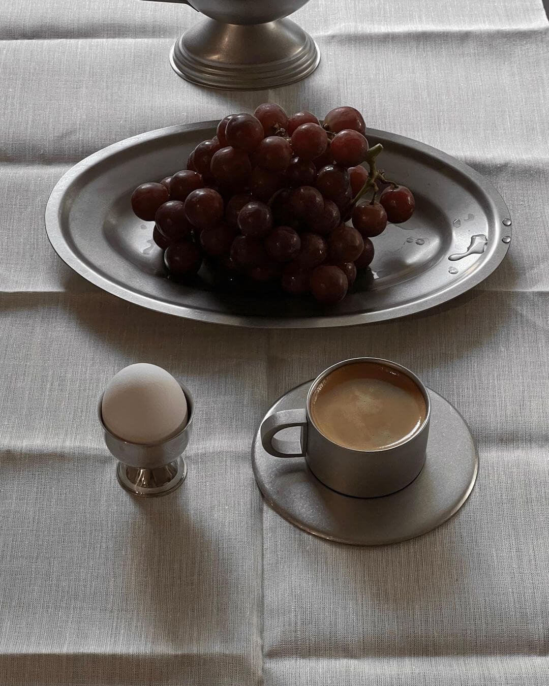 Stainless steel cup and saucer set with coffee, alongside a plate of grapes and egg on a tablecloth.