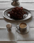 Stainless steel cup and saucer set with coffee, alongside a plate of grapes and egg on a tablecloth.