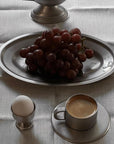 Stainless steel oval plate with grapes, egg holder, and coffee cup on a linen tablecloth.