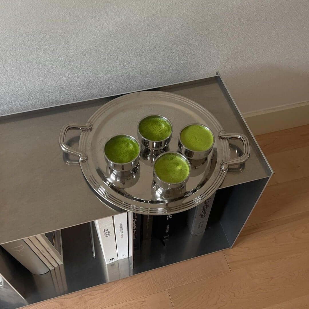 Shiny round tray holding four green drinks in metallic cups on a modern shelf with books.