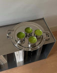 Shiny round tray holding four green drinks in metallic cups on a modern shelf with books.