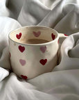 Beige hand-painted coffee mug with red and pink hearts, 300ml capacity, resting on white fabric blanket.